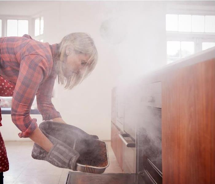 Woman taking some burnt food out of an oven in a smoke-filled kitchen.