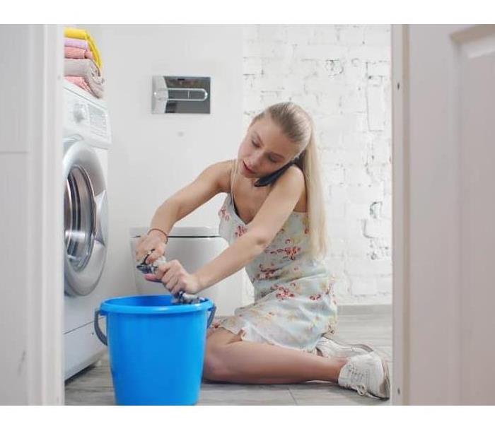 A woman cleans the floor surrounding her leaking washing machine while calling for help on her smartphone.